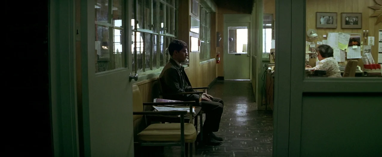 The Yards (2000) movie still: wide — A linoleum-floored corridor features wood-paneled walls and a series of glass partitions. A man waits in a…