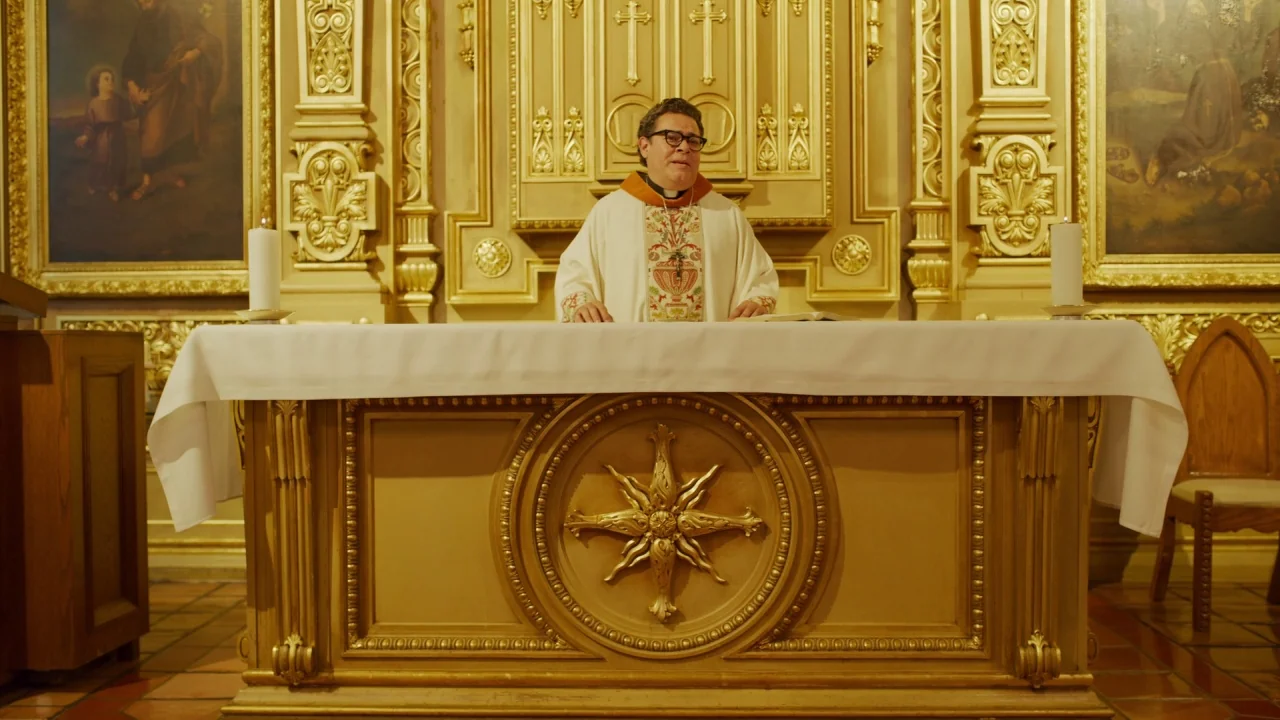 The Laundromat (2019) movie still: medium — A man in clerical vestments stands centered behind a large, ornate wooden altar. Ornate gold paneling…