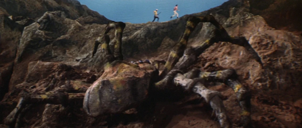 Son of Godzilla (1967) movie still: wide — A giant, monstrous arachnid creature dominates the foreground of a rugged, rocky cliffside path. Two men…