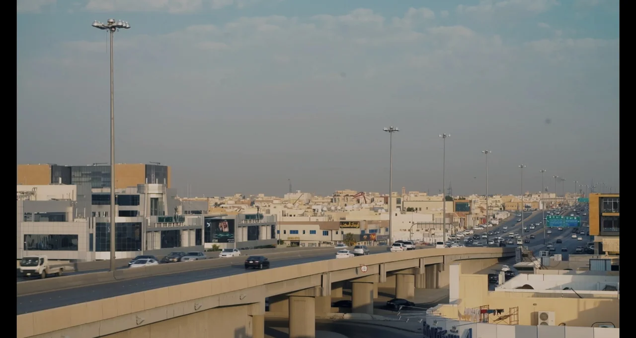 Saudi Women's Driving School (2019) movie still: establishing — An elevated concrete highway cuts through a dense cityscape under a hazy daytime…