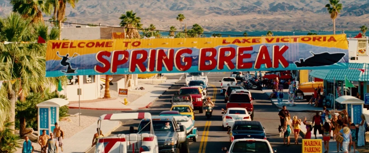 Piranha 3D (2010) movie still: establishing — A sunny lakefront street features a large banner greeting visitors for spring break festivities.…