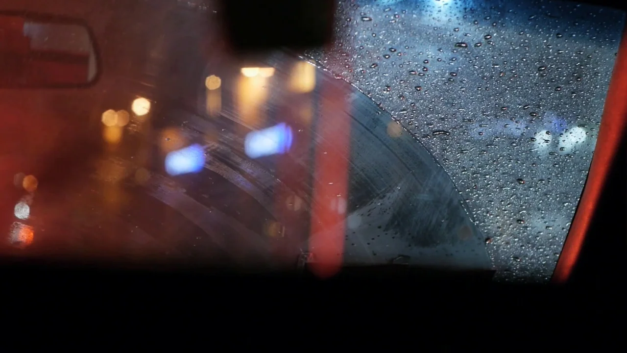 Counting (2015) movie still: closeup — A view through a wet car windshield reveals blurred city lights at night. A wiper blade trace marks a curved…