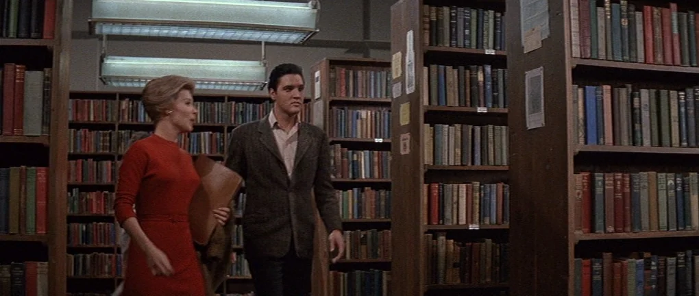 Wild in the Country (1961) movie still: medium — Rows of tall wooden shelves packed with colorful book spines fill the indoor space. A man and woman…