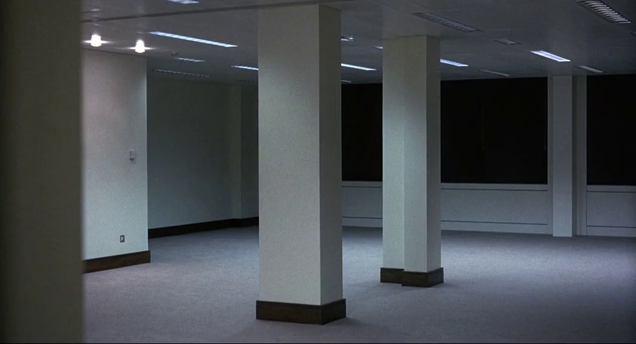 Naked (1993) movie still: wide — A vacant corporate floor features rows of white pillars and grey carpeted flooring. Large dark windows line the…