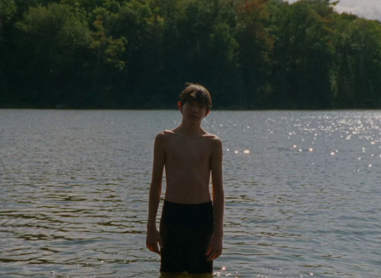Falcon Lake (2022) movie still: fullbody — A calm lake surrounded by a dense green forest under bright daylight. A young man stands waist-deep in…