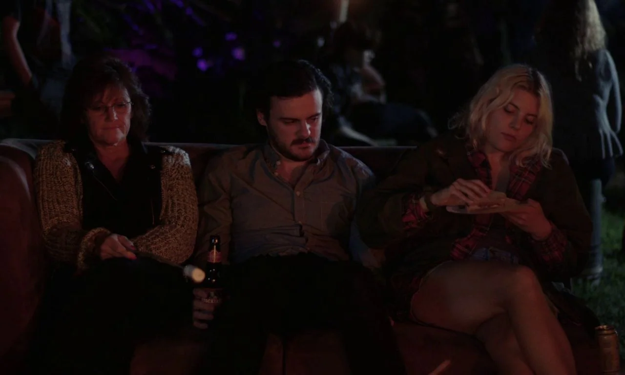 Ham on Rye (2019) movie still: medium — Three people sit together on a dark couch at night during an outdoor gathering. The woman on the right holds…