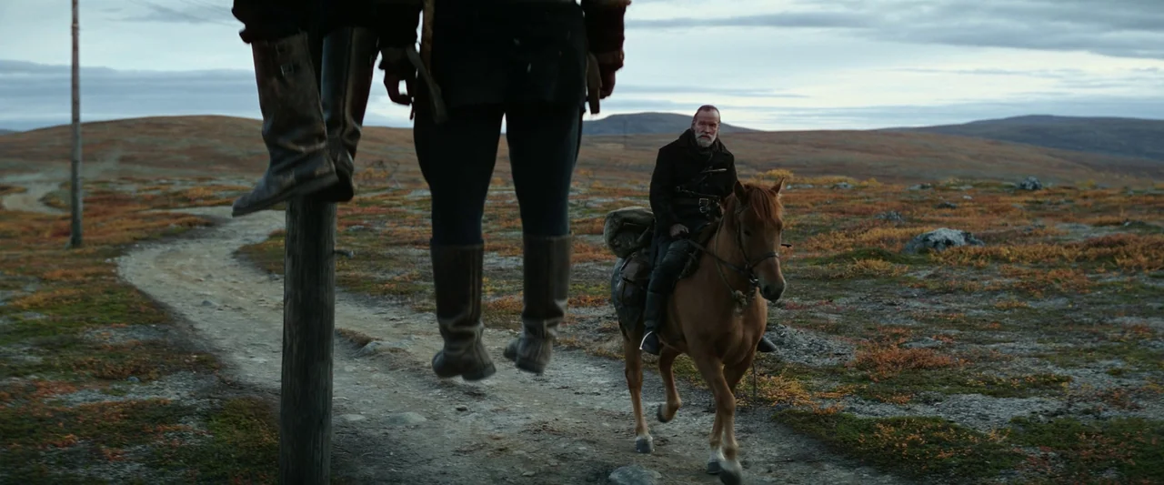 Sisu (2022) movie still: medium — A desolate tundra landscape features a dirt path under an overcast sky. A man rides a brown horse past hanging…
