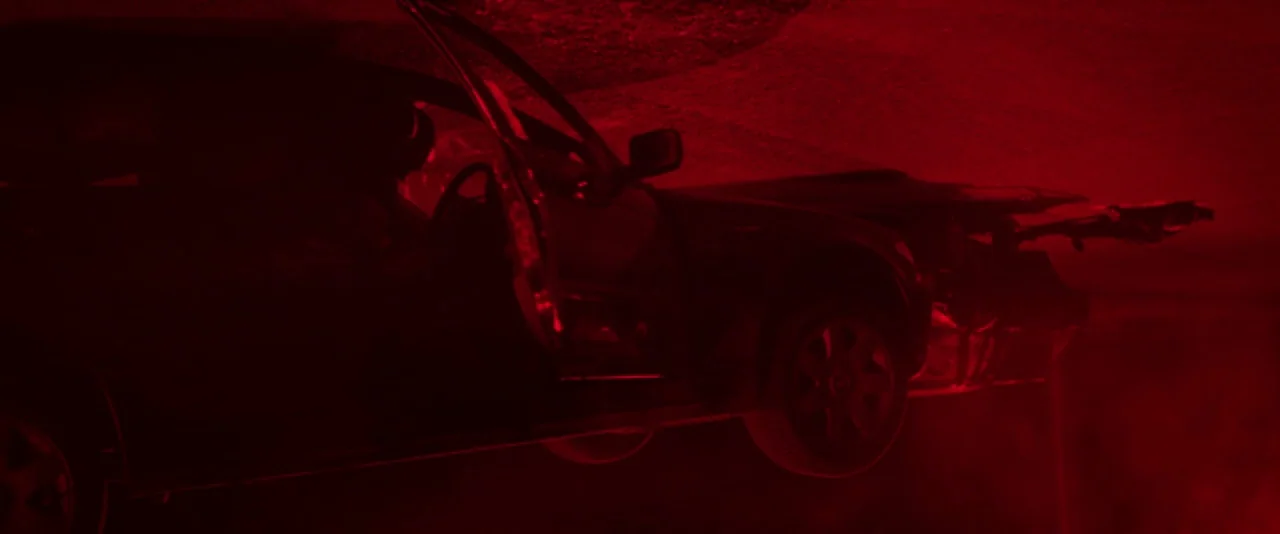 Kissing Candice (2017) movie still: medium — A damaged passenger car is positioned on an asphalt surface under dark conditions. The front bumper and…