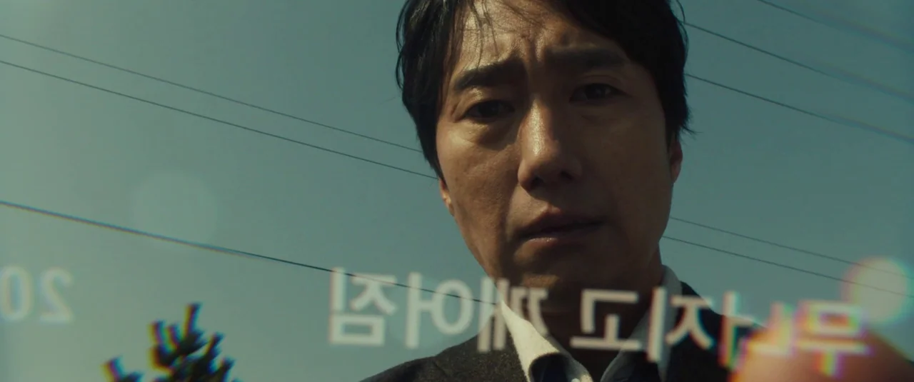Decision To Leave (2022) movie still: closeup — A man looks down at the camera against a clear blue sky. Thin black power lines cross the frame…