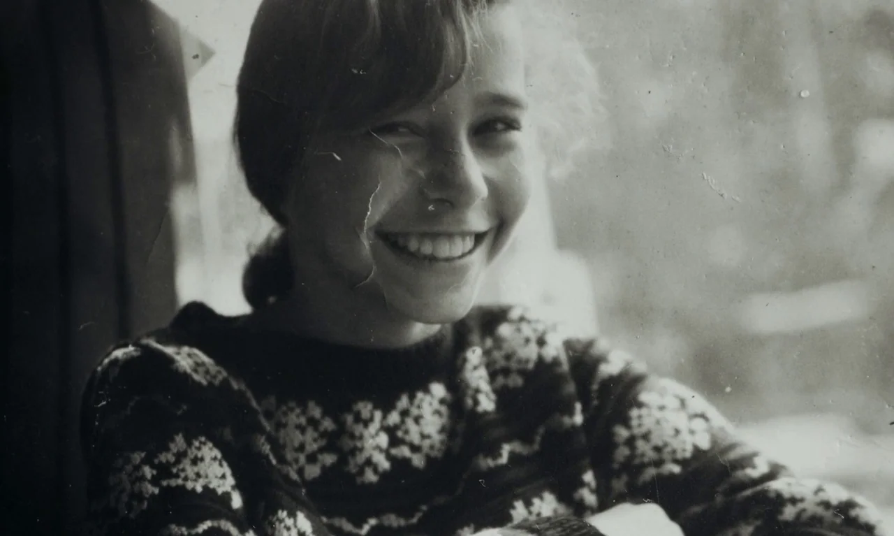 Little Girl Blue (2023) movie still: closeup — A portrait of a young woman in a vehicle with a window visible behind. She smiles directly at the…
