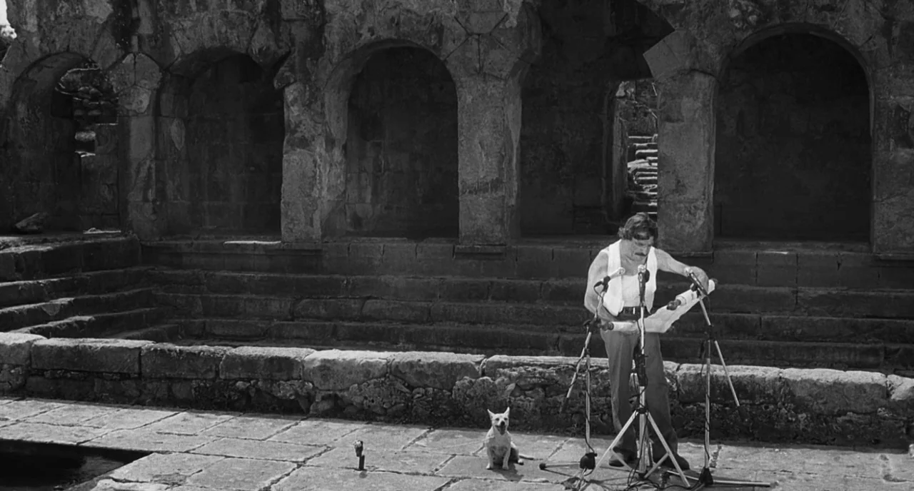 The Legend Of Kaspar Hauser (2012) movie still: wide — A man stands before three microphones in the courtyard of a weathered stone ruin. He reads…