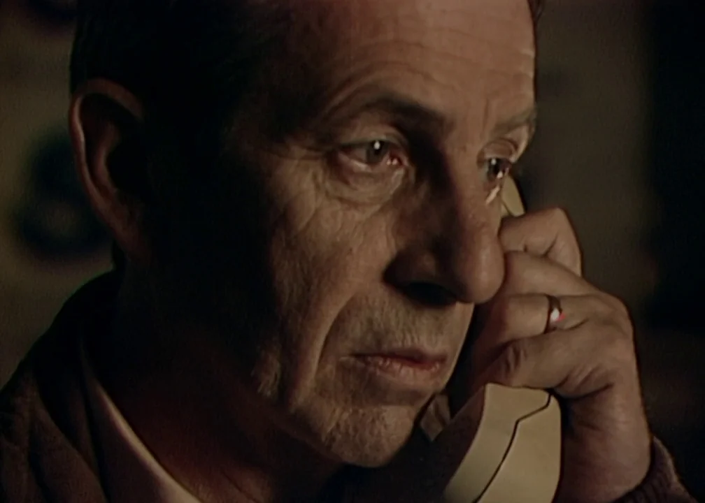 No (2012) movie still: closeup — An interior space is dimly lit, focusing on an elderly man's face. He holds a beige telephone handset against his…