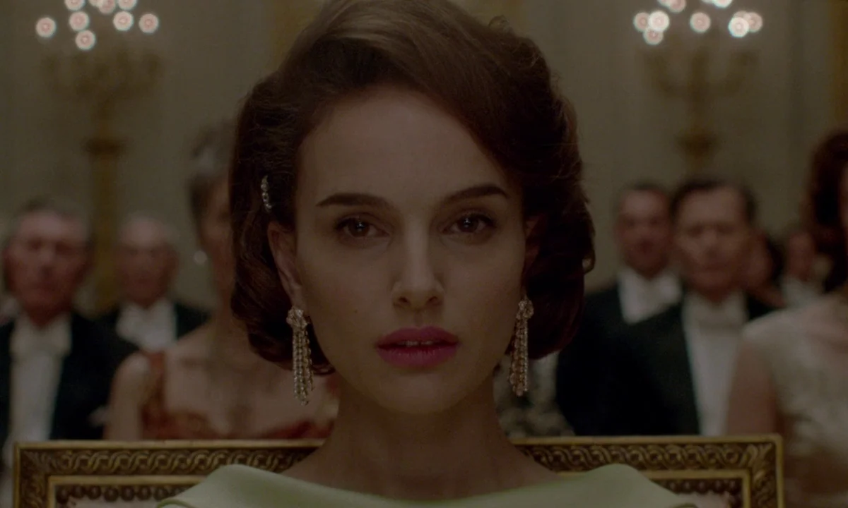 Jackie (2016) movie still: closeup — A formal ballroom filled with guests in evening wear seated in ornate gold chairs. A woman looks directly…