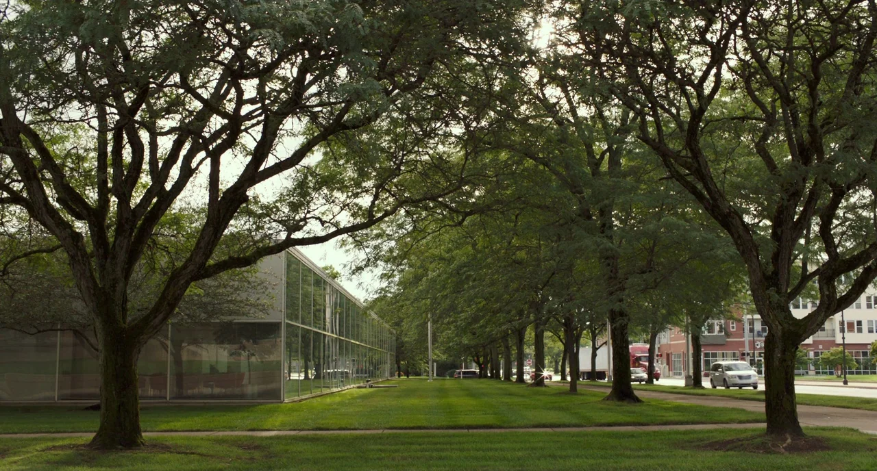 Columbus (2017) movie still: establishing — A well-manicured lawn is surrounded by large trees and a modernist glass building facade. A road with a…