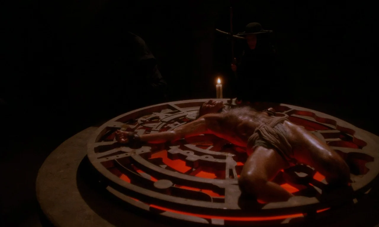 The Church (1989) movie still: wide — A dark, dimly lit stone chamber contains a large, circular, gear-like metallic structure. A shirtless man is…