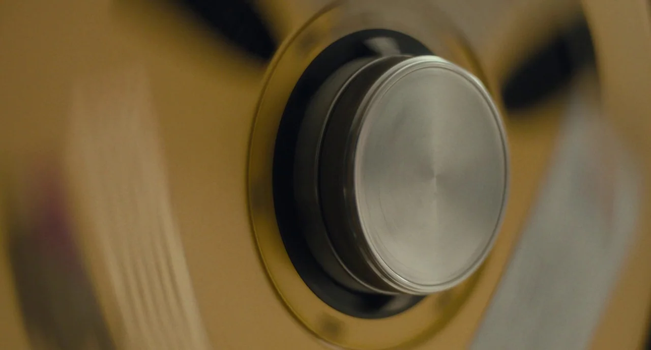 Christine (2016) movie still: closeup — A close-up shot of a vintage gold-colored magnetic tape reel hub. The center circular plate is made of…