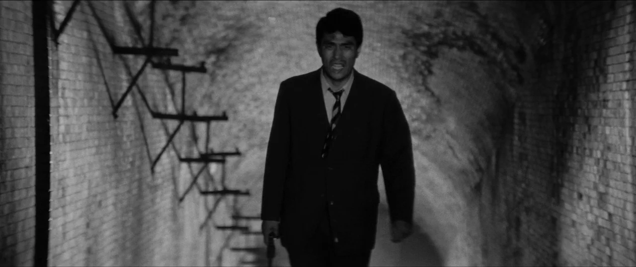 Branded To Kill (1967) movie still: medium — A narrow tunnel features curved brick walls with metal rungs attached to the side. A man walks toward…
