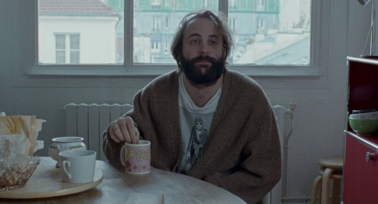 Non-Fiction (2018) movie still: medium — A man sits at a round wooden table in a room with large windows. He holds a ceramic mug while looking ahead…