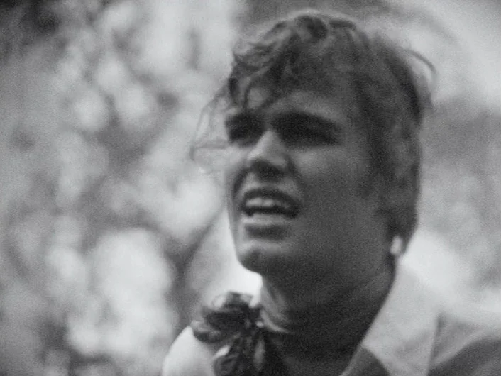 Eika Katappa (1969) movie still: closeup — An outdoor setting with out-of-focus foliage visible in the background. A young man with wavy hair looks…