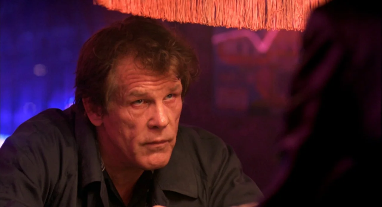 The Good Thief (2002) movie still: closeup — A dimly lit interior space with a fringed lampshade overhead and red lighting. A man with graying brown…