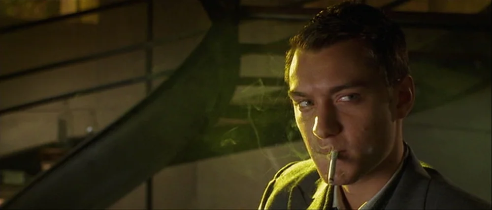 Gattaca (1997) movie still: closeup — A man stands near the metal railing of a dark indoor staircase. He smokes a cigarette while facing the camera…