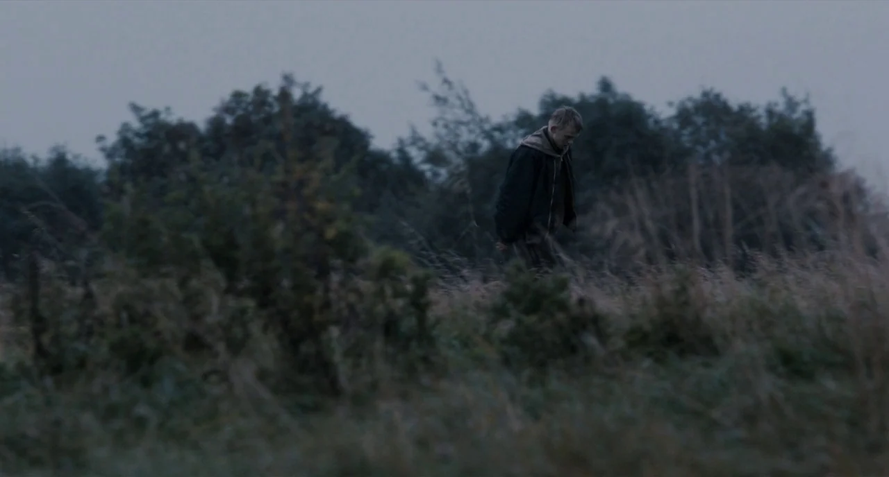 The Selfish Giant (2013) movie still: wide — An open field with tall, dried grasses under an overcast, grey sky. A young man walks away from the…