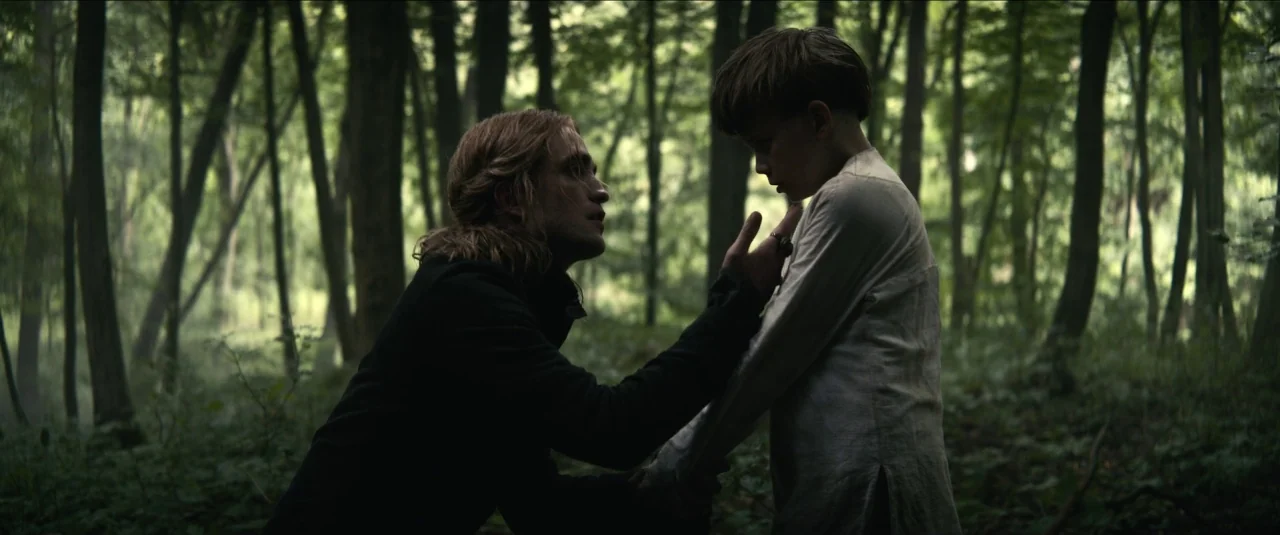 The King movie still: medium — Densely packed trees and undergrowth form a dark, shadowed clearing in the woods. A kneeling man reaches out to…