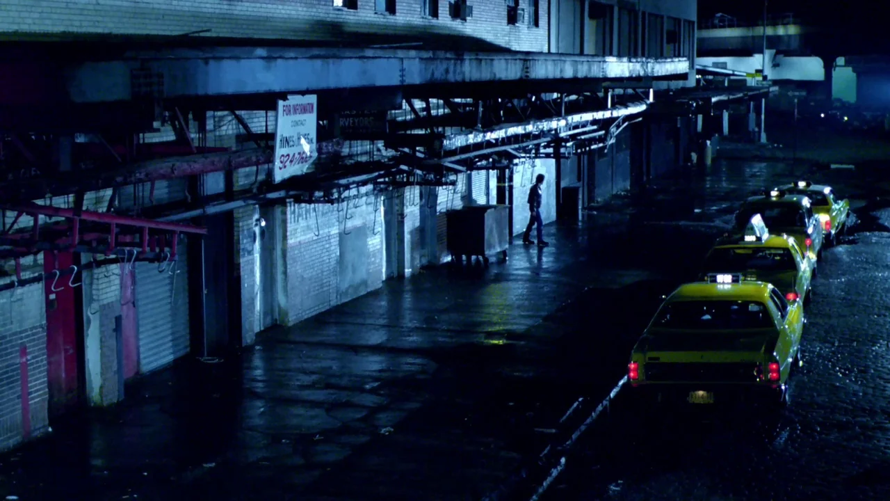 Cruising (1980) movie still: wide — A dark urban alleyway features brick structures and a long row of yellow taxis. A man walks slowly past the…