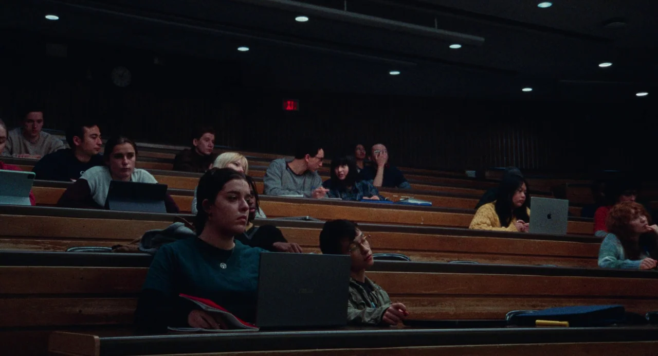 Dream Scenario (2023) movie still: wide — A dimly lit university lecture hall with tiered wooden seating rows filled with students. Students sit at…