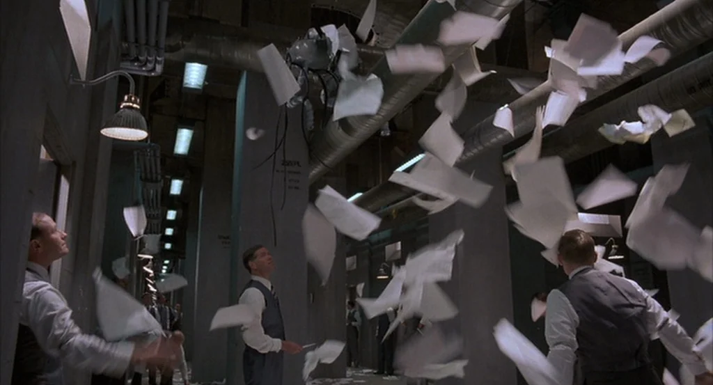Brazil (1985) movie still: wide — An industrial office space features exposed overhead metal ductwork and concrete support pillars. Several men in…