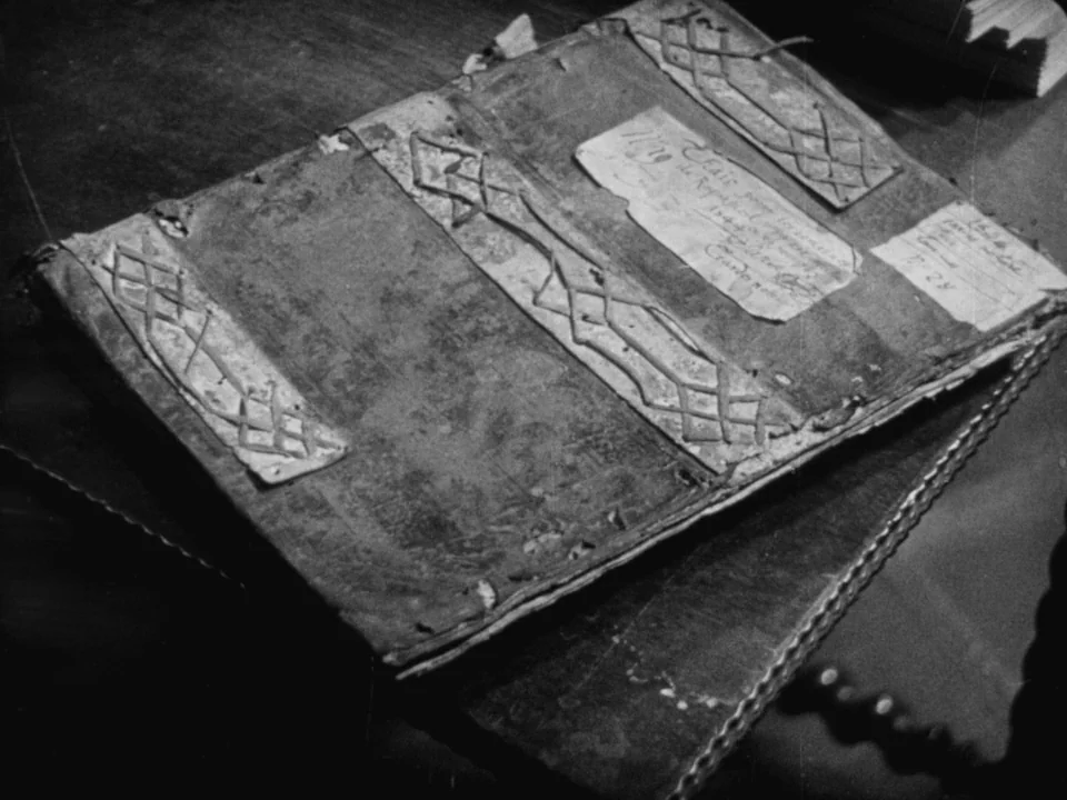 The Passion of Joan of Arc (1928) movie still: closeup — An old worn ledger rests on a dark table surface under soft lighting. The antique book…