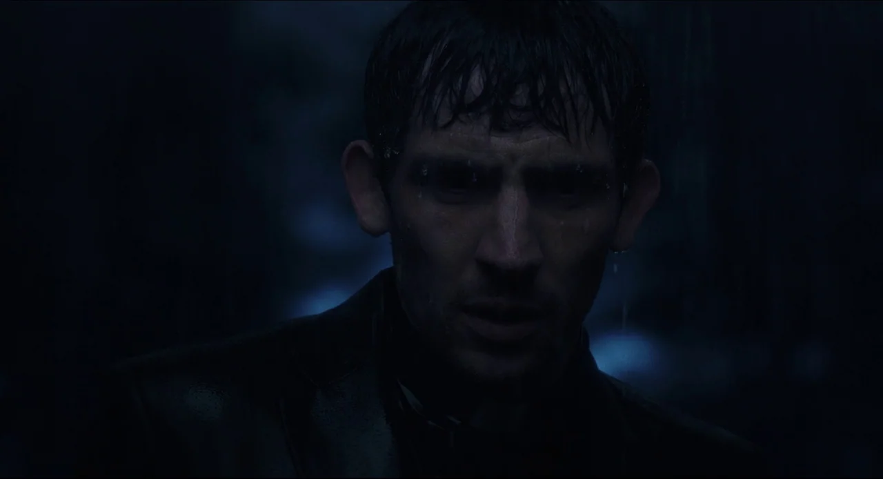 Wake Up Dead Man (2025) movie still: closeup — A man stands outdoors at night during a rainstorm. Raindrops glisten on his dark, saturated hair and…