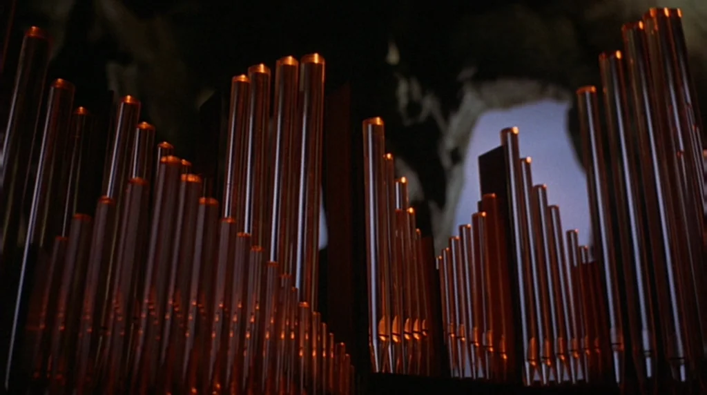 Danger Diabolik (1968) movie still: closeup — A close view of metallic organ pipes arranged in height-based rows. Dark shadows frame the pipes…