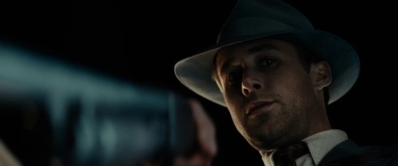 Gangster Squad (2013) movie still: closeup — A dark night setting with minimal background detail visible behind the man. A man wearing a fedora…