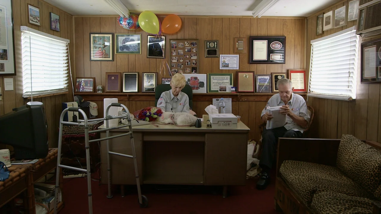 The Last Race (2022) movie still: wide — An office with wood-paneled walls decorated with framed certificates, photos, and colorful balloons. An…