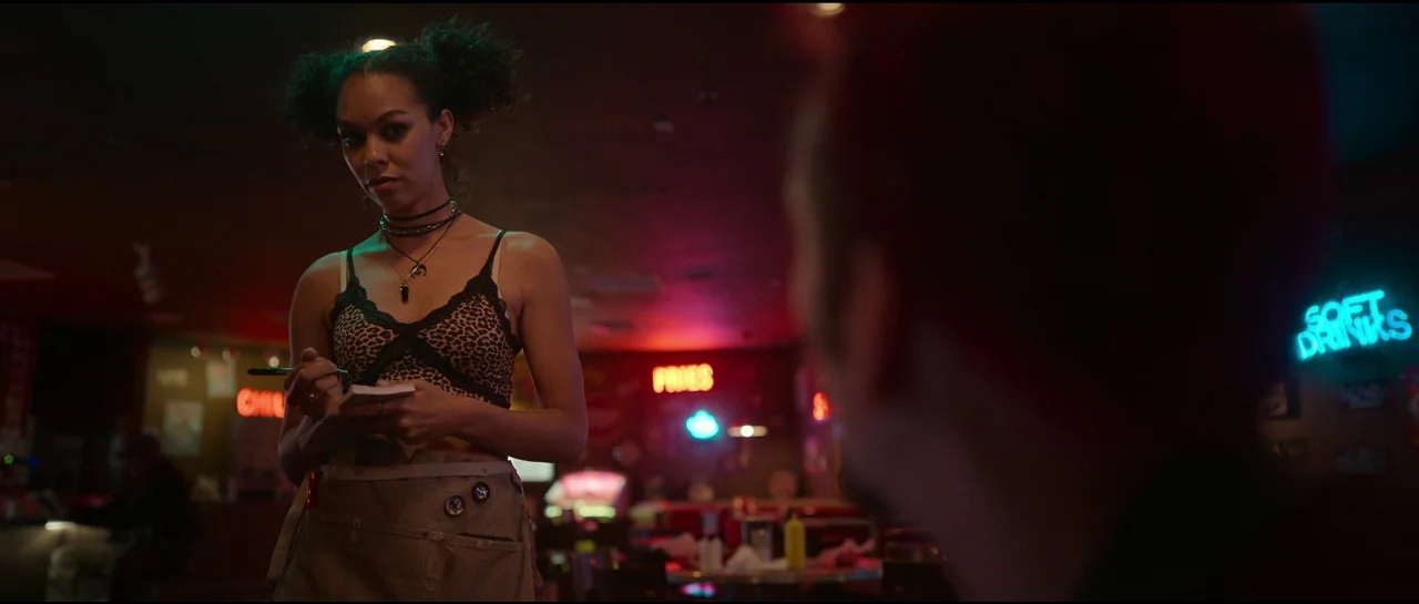 Sympathy For The Devil (2023) movie still: medium — A dimly lit restaurant interior with vibrant neon signs on the dark walls. A waitress stands…