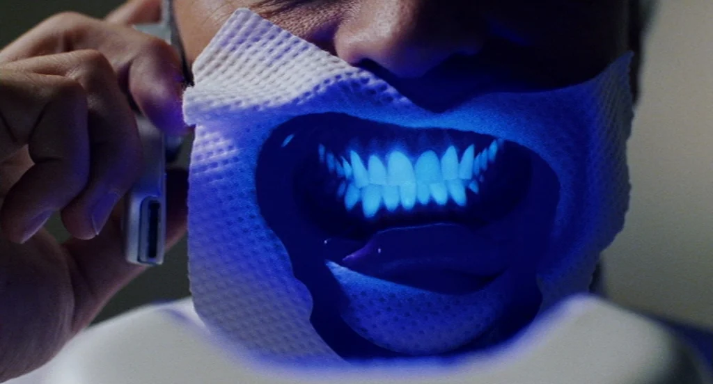Intolerable Cruelty (2003) movie still: closeup — The frame shows a close-up of a person wearing a dental bib. They hold a white smartphone against…