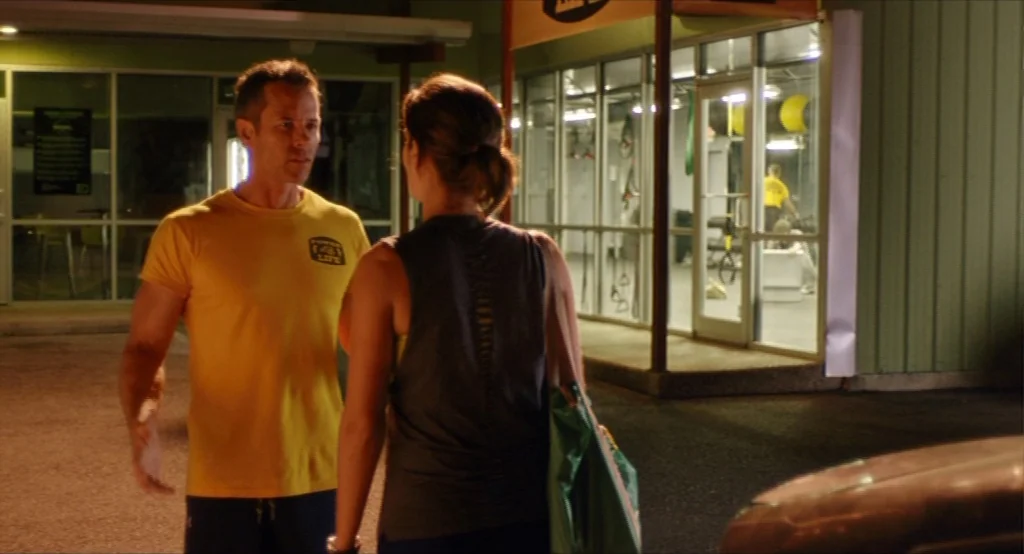 Results (2015) movie still: medium — A dark outdoor parking lot at night adjacent to a brightly lit commercial building. A man in a yellow t-shirt…