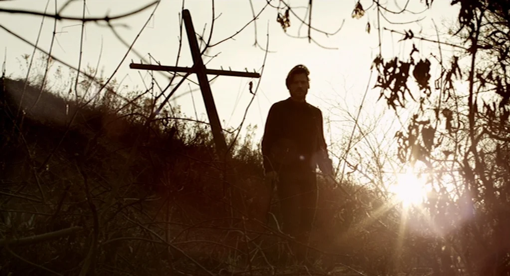 Mama (2013) movie still: fullbody — An overgrown area with dry, leafless branches and tall grass surrounds a person. A wooden cross stands fixed in…