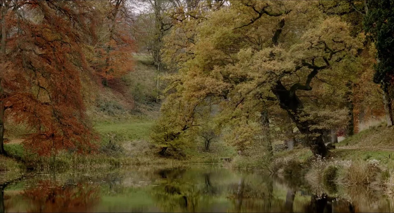 The Falling movie still: establishing — A calm, reflective pond is surrounded by dense trees with orange and green foliage. The water reflects the…