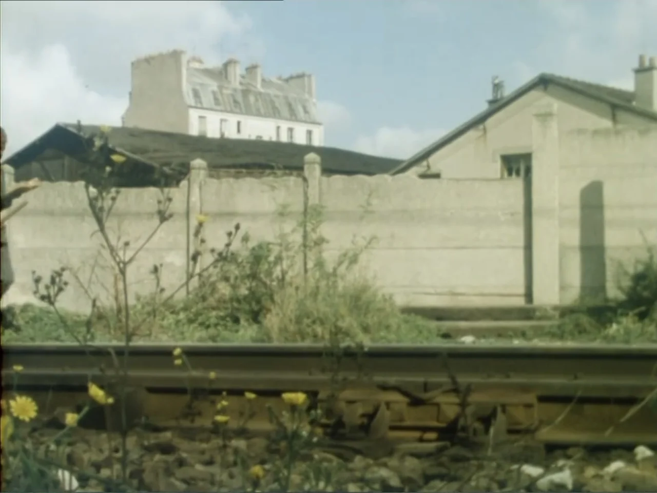 A Dessert for Constance (1981) movie still: wide — A quiet exterior scene next to train tracks with a beige concrete wall behind. A young man stands…