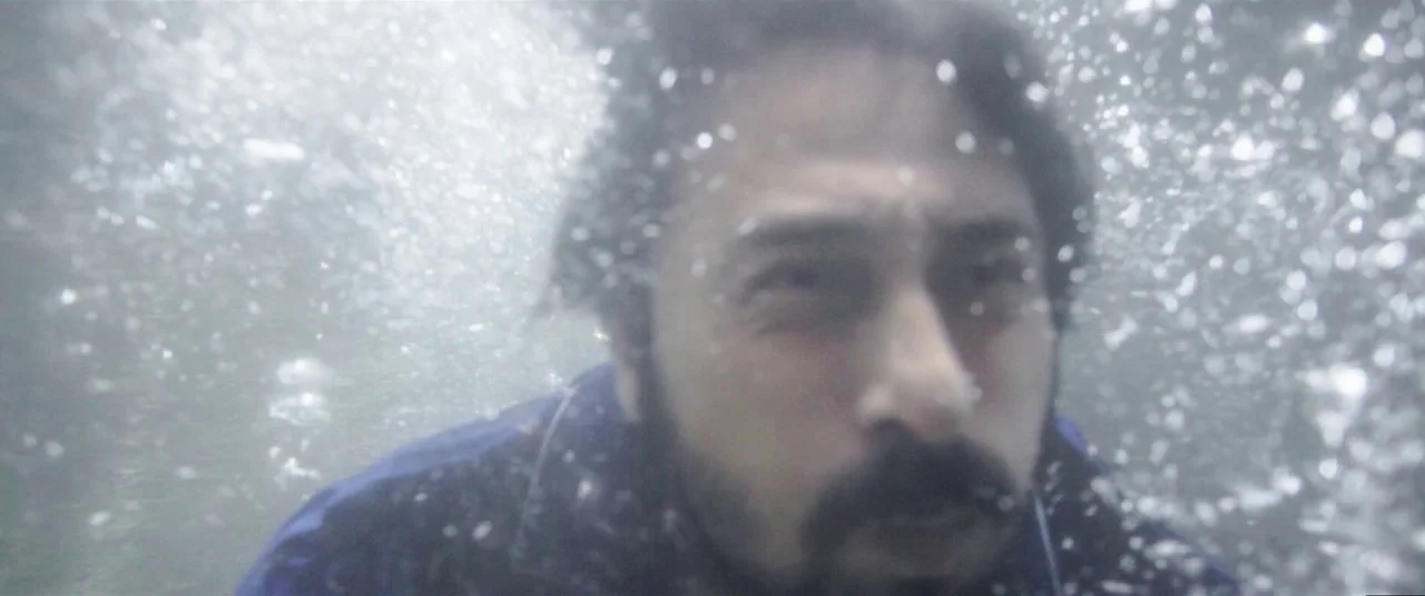 Satan’s Slaves (2017) movie still: closeup — A man is submerged underwater with a high volume of air bubbles surrounding him. He stares directly at…