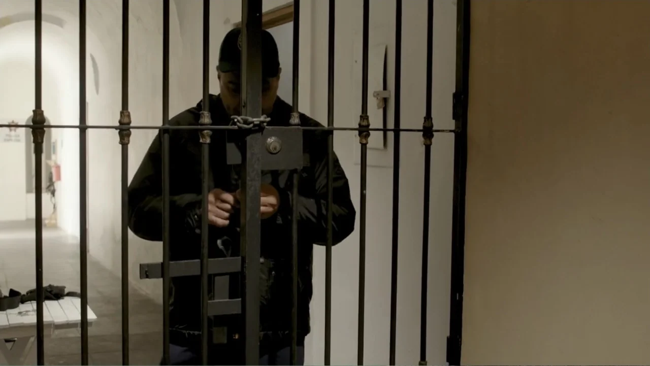 Por Amor al Money (2023) movie still: medium — A man stands behind the iron bars of a secure gateway in a hallway. The man handles a key or metallic…