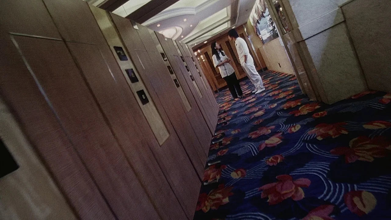 Visible Secret (2001) movie still: wide — A long hotel corridor features rows of wooden paneled doors and patterned floral carpet. A man and a woman…