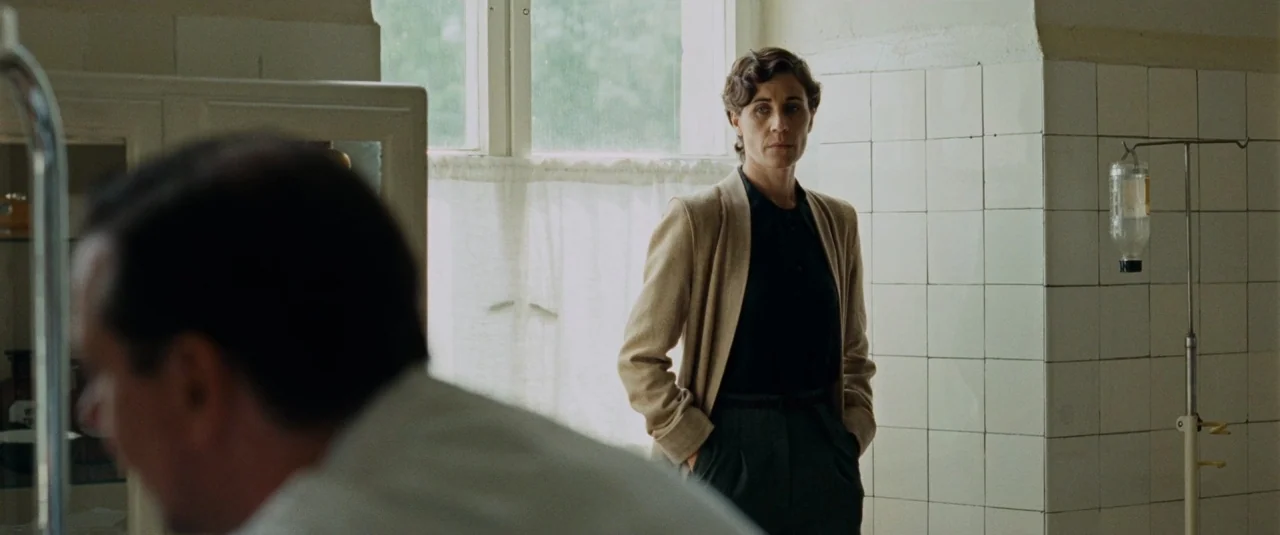 Phoenix (2014) movie still: medium — A sterile clinical room features white tiled walls and a large, bright window. A woman with short wavy hair…