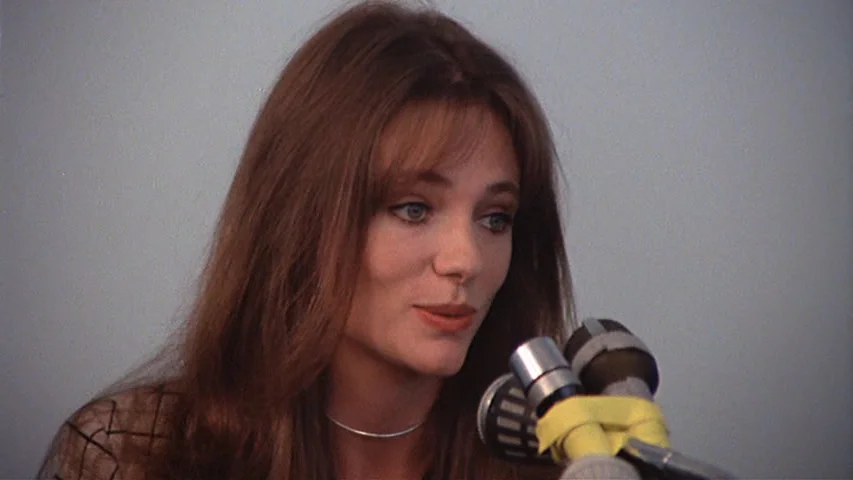 Day for Night (1973) movie still: closeup — A woman stands before a pale gray wall during a press interview setup. She speaks into a dual-microphone…
