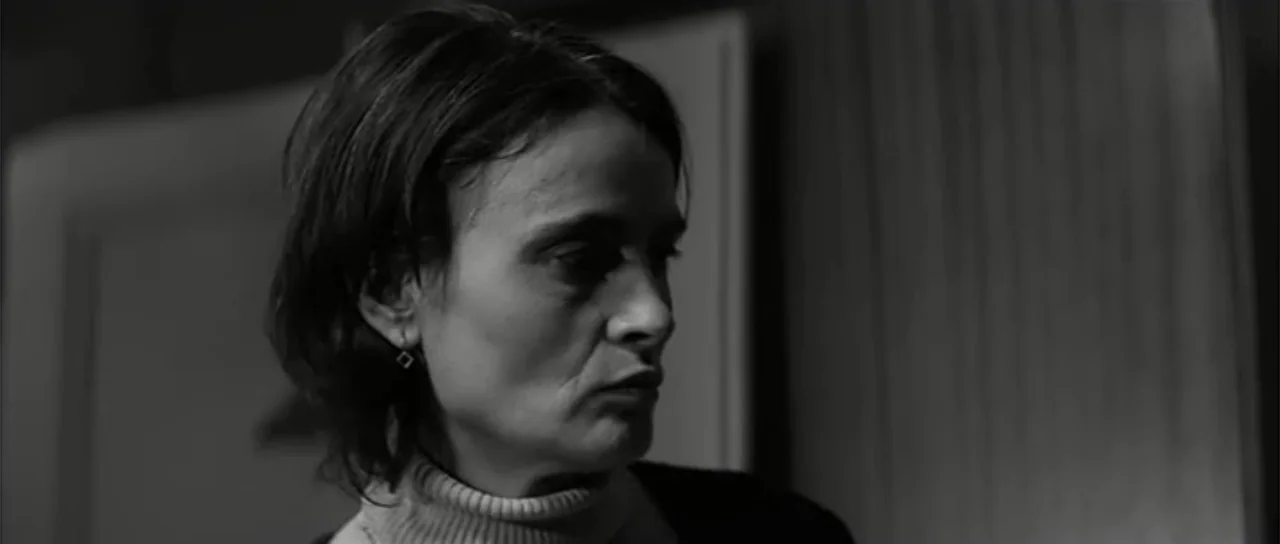 13 Tzameti (2006) movie still: closeup — A dark indoor space with minimal background elements and plain surfaces visible. A woman in a turtleneck…