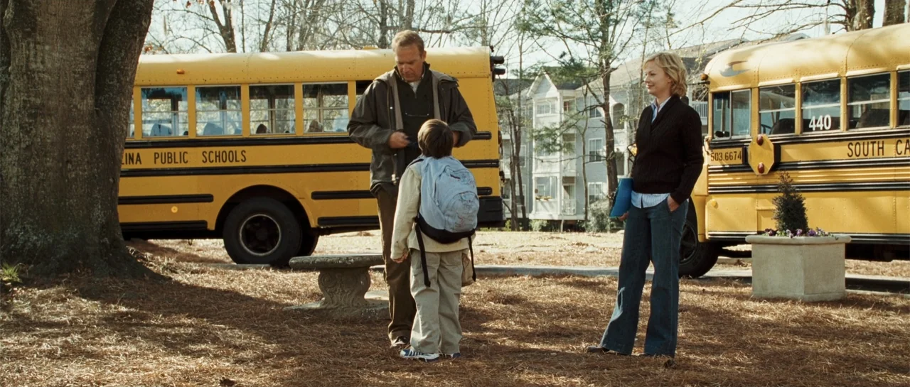 The New Daughter (2009) movie still: fullbody — A suburban school parking lot with two large yellow buses parked in the background. A man, a boy,…