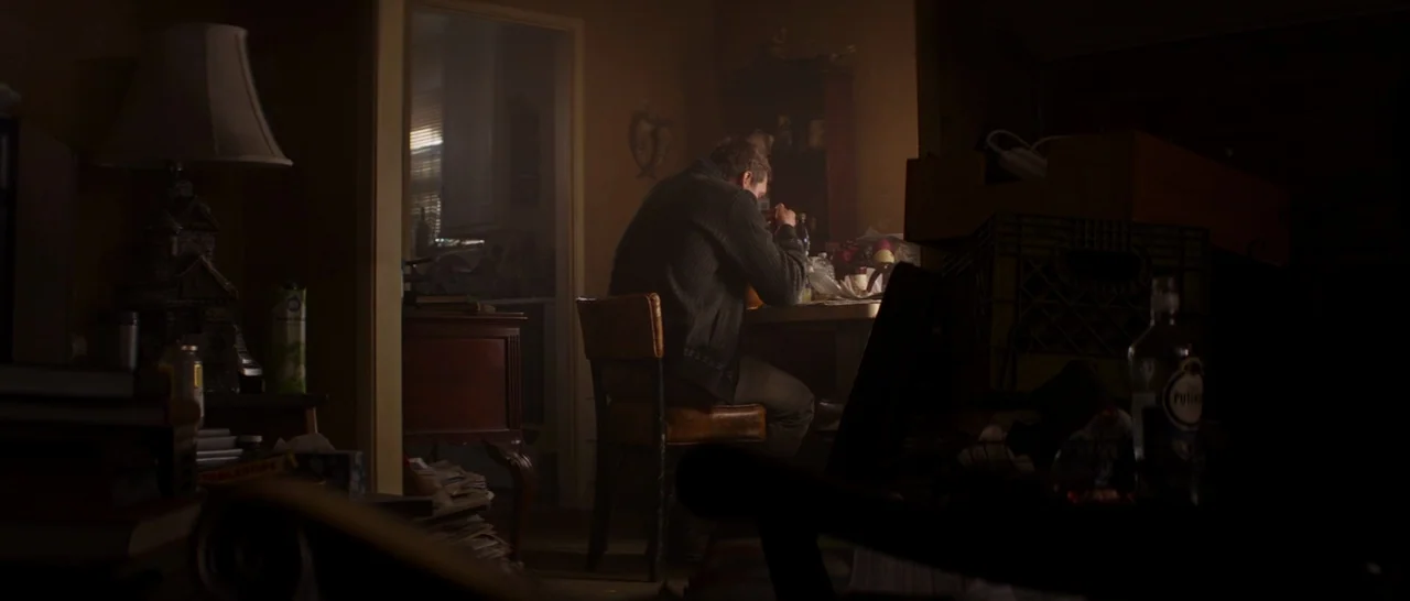 Captain America: Civil War (2016) movie still: medium — A dimly lit room contains a cluttered dining table and stacks of old newspapers. A man sits…