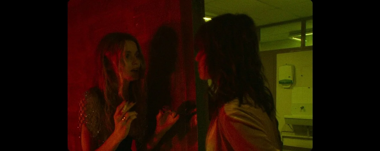 Lux Æterna (2019) movie still: medium — A public restroom with tiled walls is divided by harsh red and green light. Two women stand across from each…